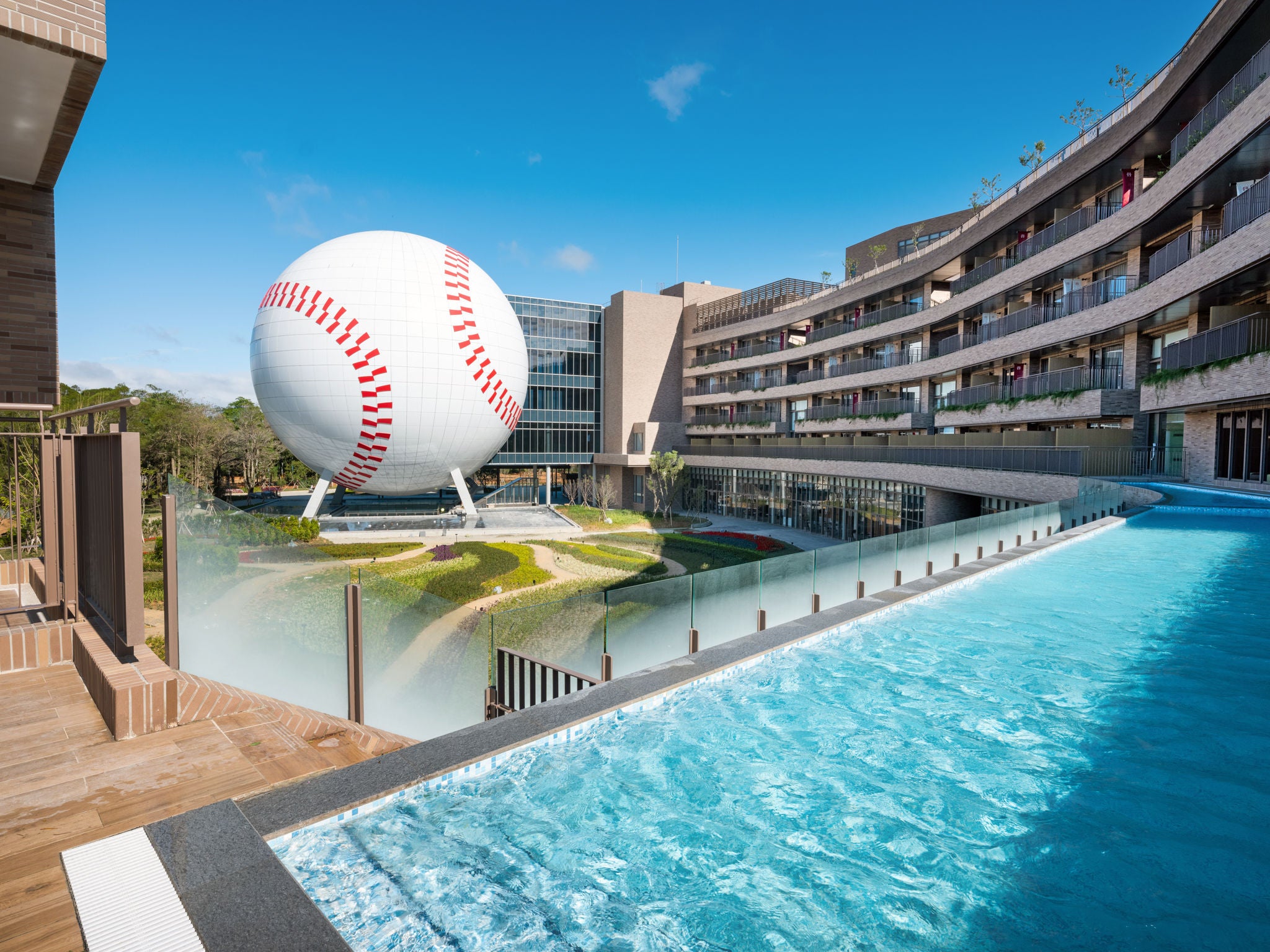An outdoor hotel swimming pool beside a curved building, with a giant baseball sculpture under a clear blue sky.