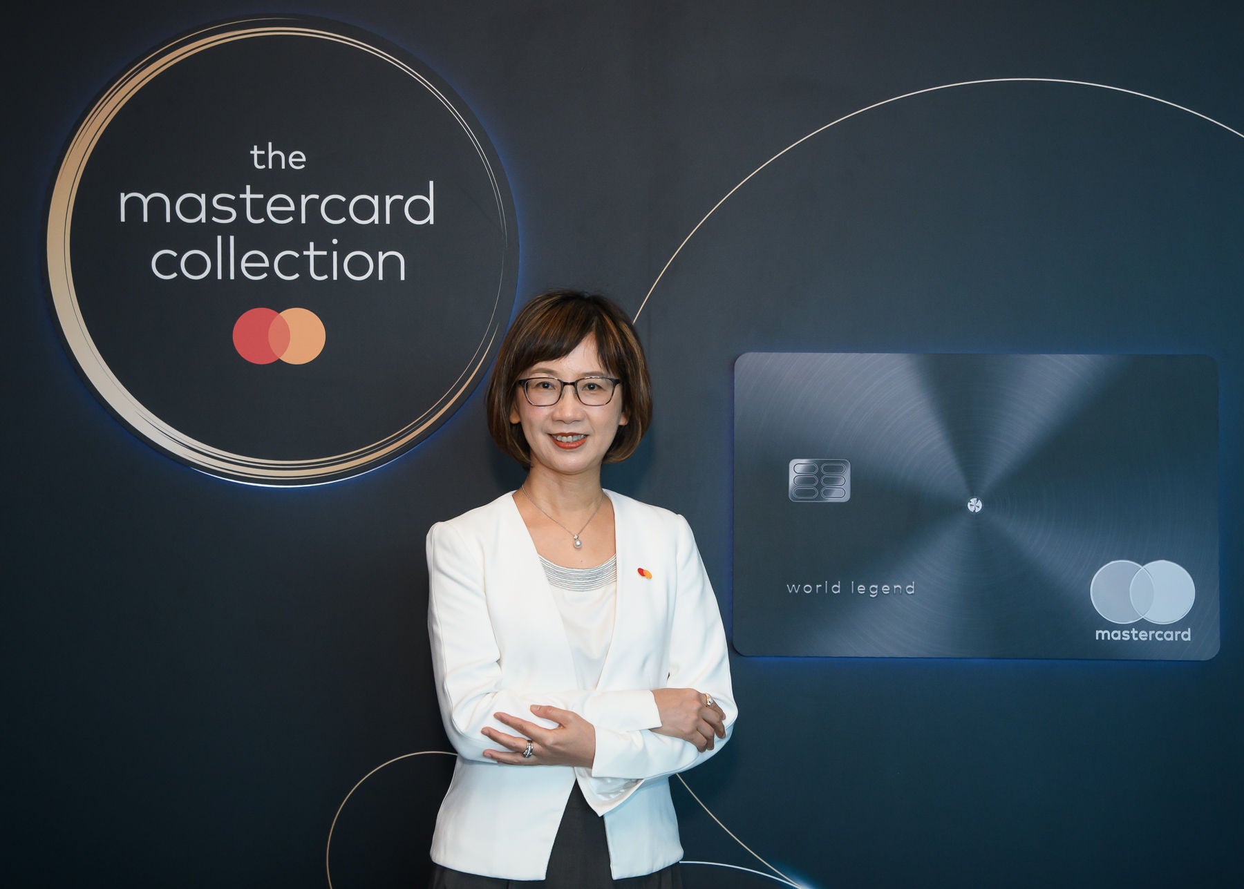 A woman in a white blazer stands with her arms crossed, smiling in front of a dark backdrop displaying a metal payment card and a brand logo.