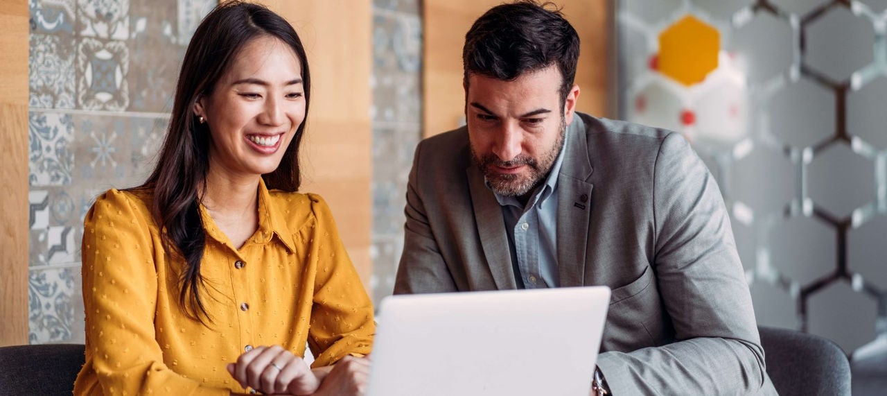 Two businesspeople look at a laptop in an office setting. 
