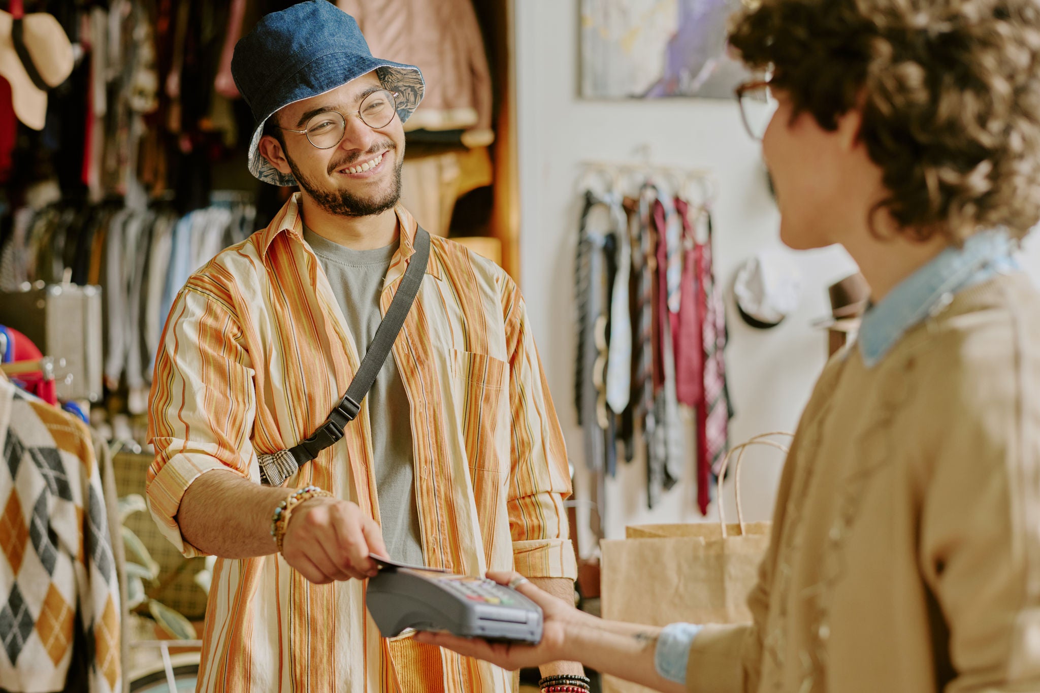 Young man wearing blue hat and glasses, smiling while making payment using card reader in a cozy clothing store with bags on table and shelves showcasing clothes around