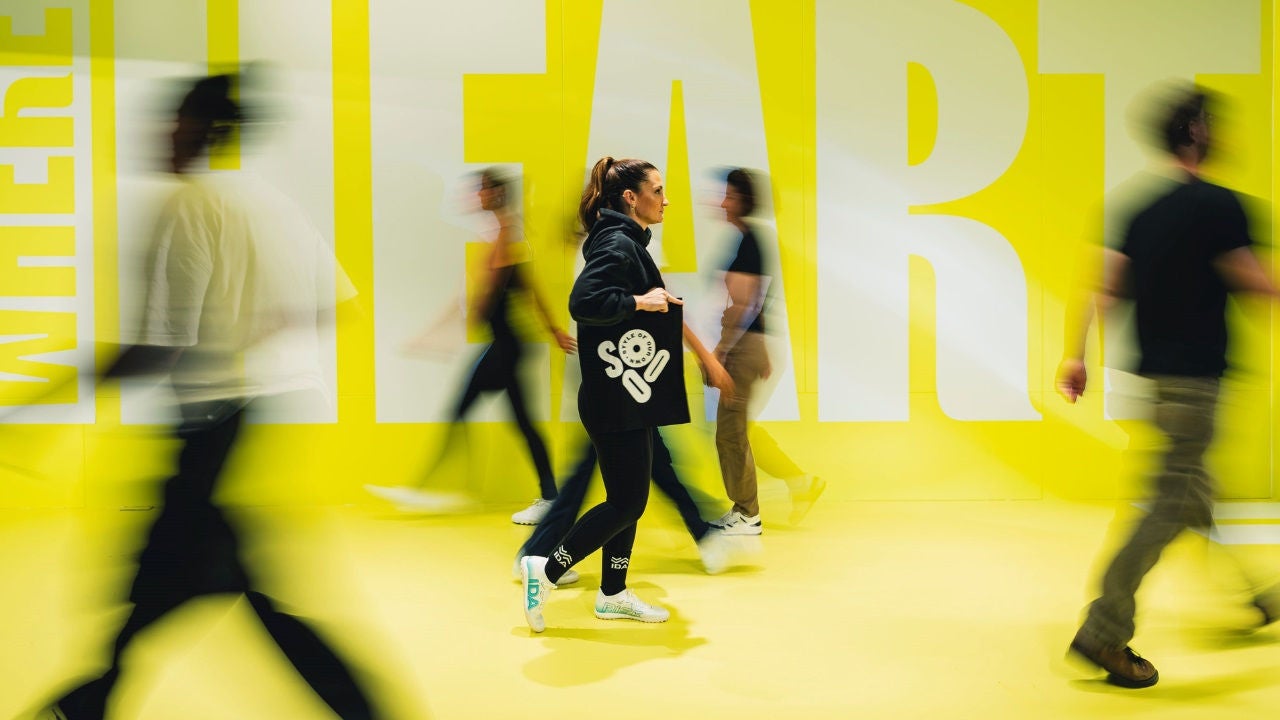 A woman in athletic wear carries an SOOO bag against a yellow background. 