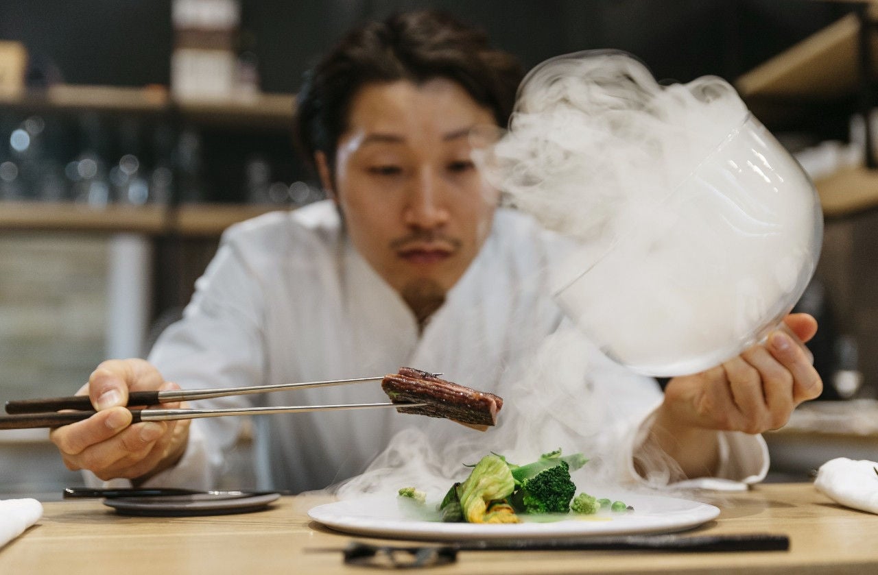 A chef holds a piece of meat over a dish while he lifts a cloque filled with steam. 
