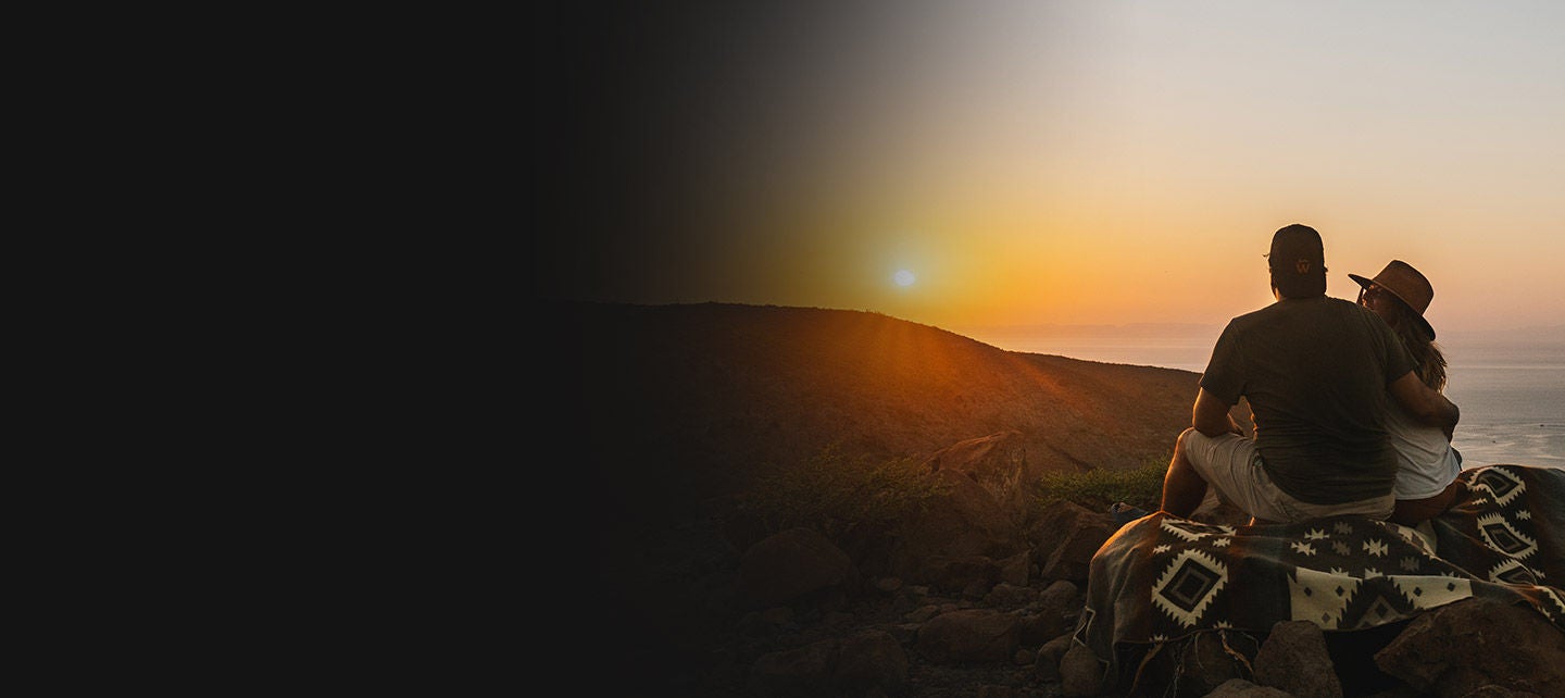 A couple sitting on a mountaintop, looking at sunset.