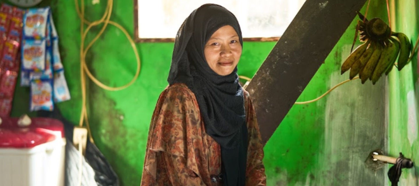 A woman in a headscarf stands in her kitchen over a stove. 