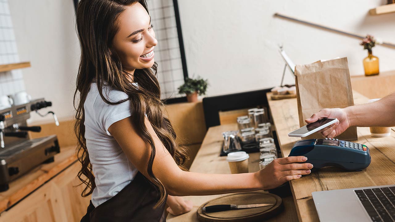 A cashier is holding terminal whilea  man is paying with smartphone in a coffee house.