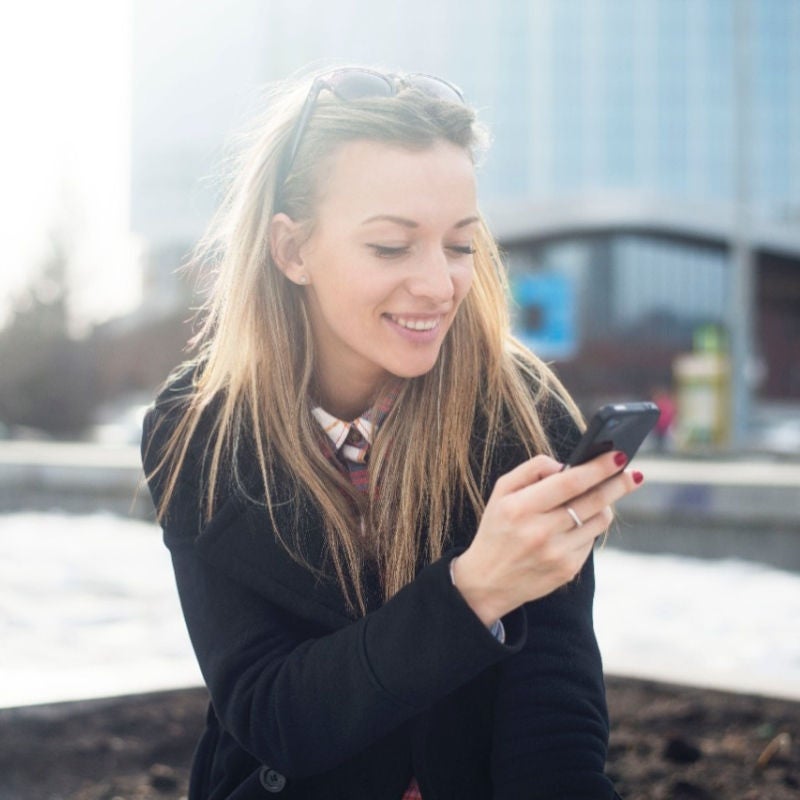 A younger woman sitting outside next to an office building looks at her smartphone.