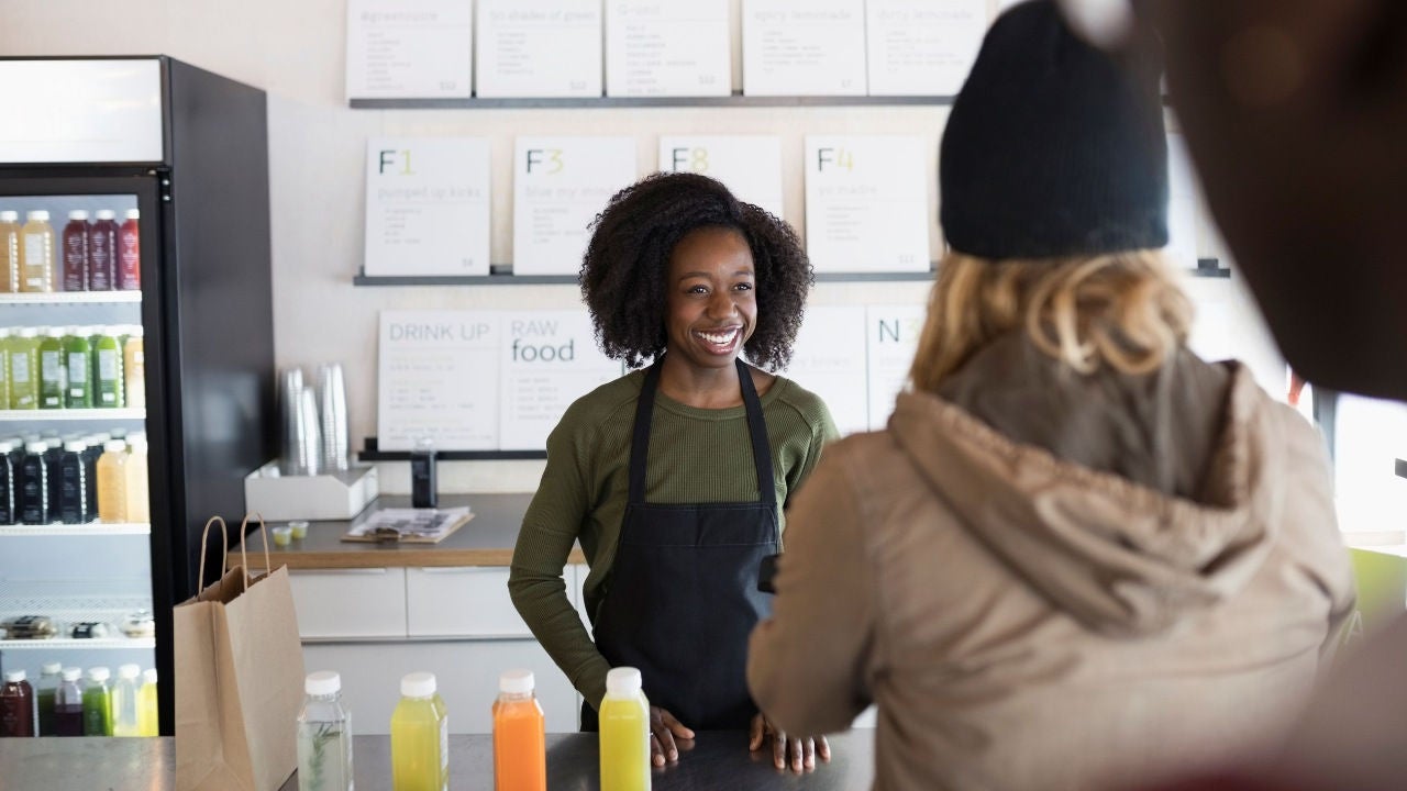 A woman in an apron at a juice bar smiles at a customer.