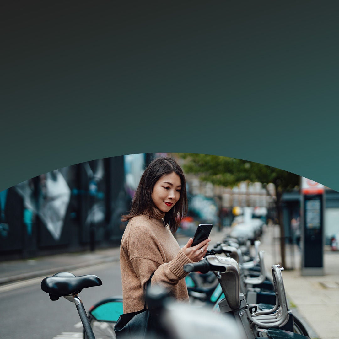 Woman looking at her phone for latest news updates near docked rental bicycles on a city street.
