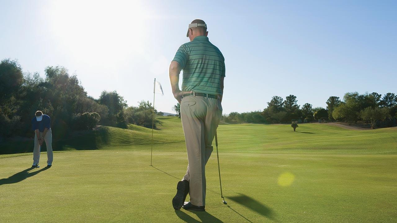 Két férfi golfozik egy napsütéses, zöld golfpályán; egyikük puttol, a másik a zászló mellett áll és nézi a játékot.