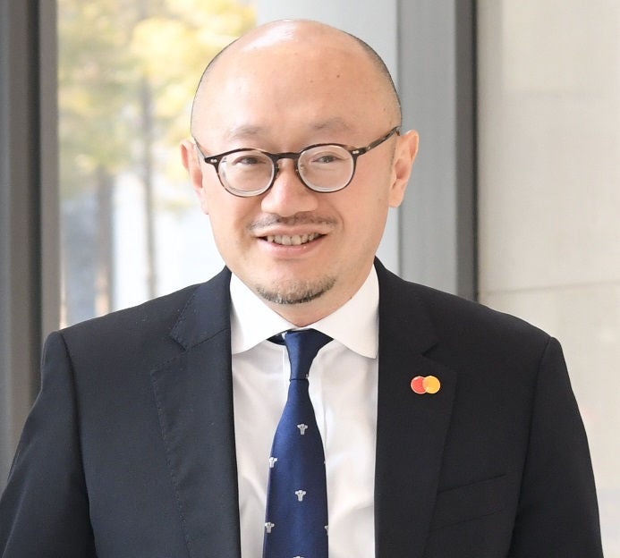 A portrait of a man in a dark suit and navy tie with glasses, smiling in front of a blurred window background.