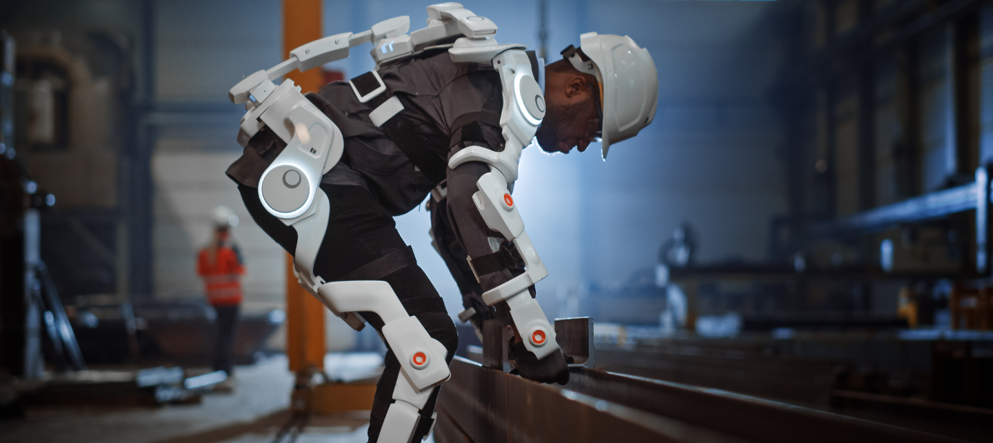 A man wearing an exoskeleten bends over to lift something in  an industrial building site.