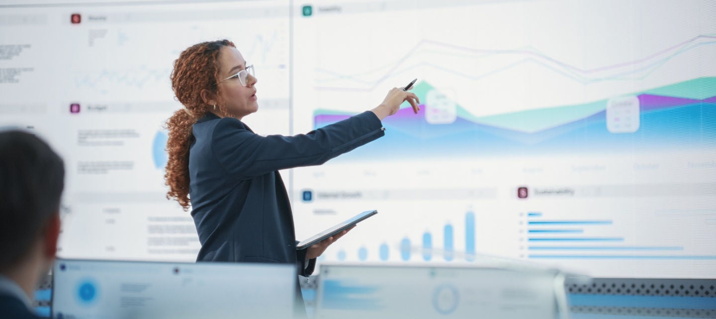 A woman in a suit jacket holds a tablet a points to a large screen showing graphics. 