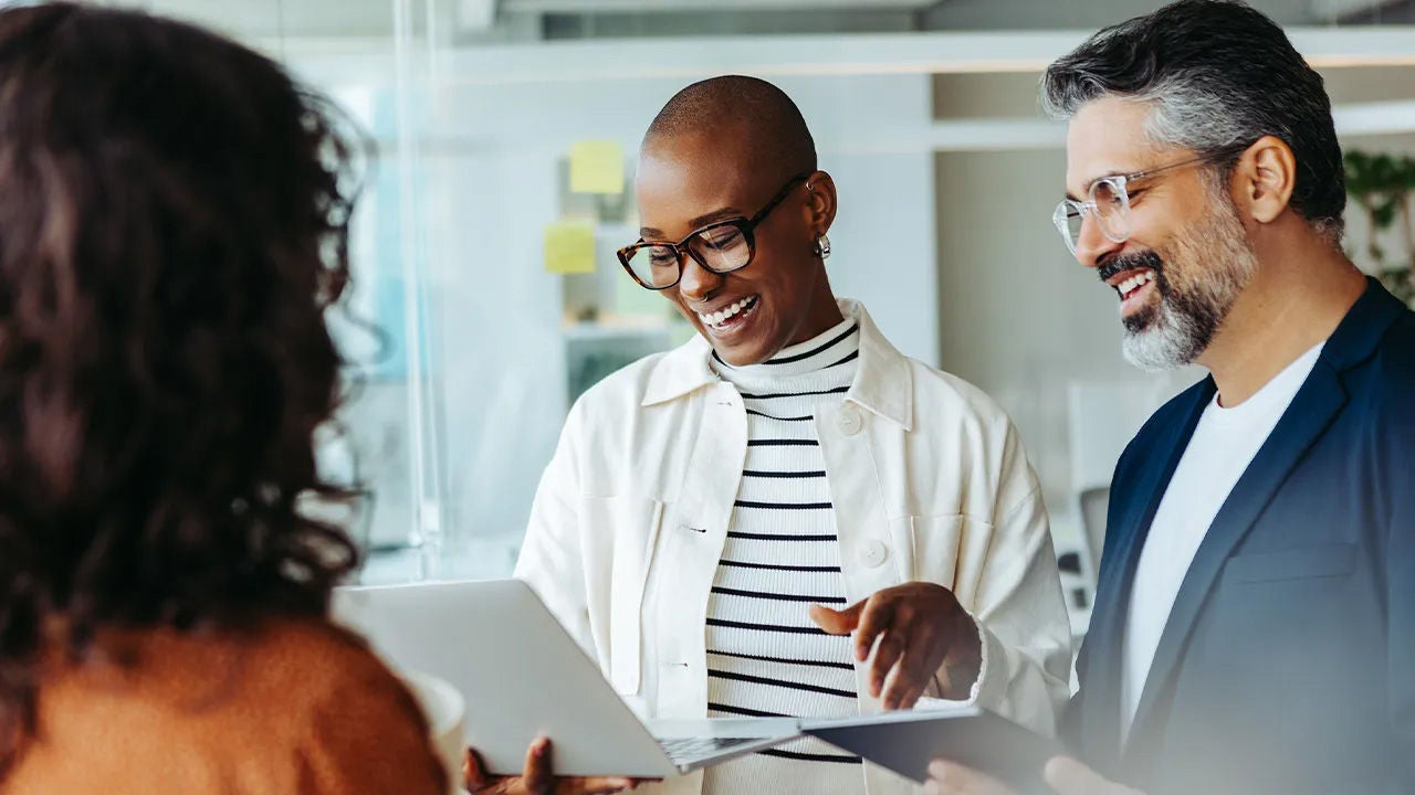 Three colleagues engaging collaboratively around digital devices within bright open-plan office environment; two hold tablets while third looks over shoulder smilingly conversing together.