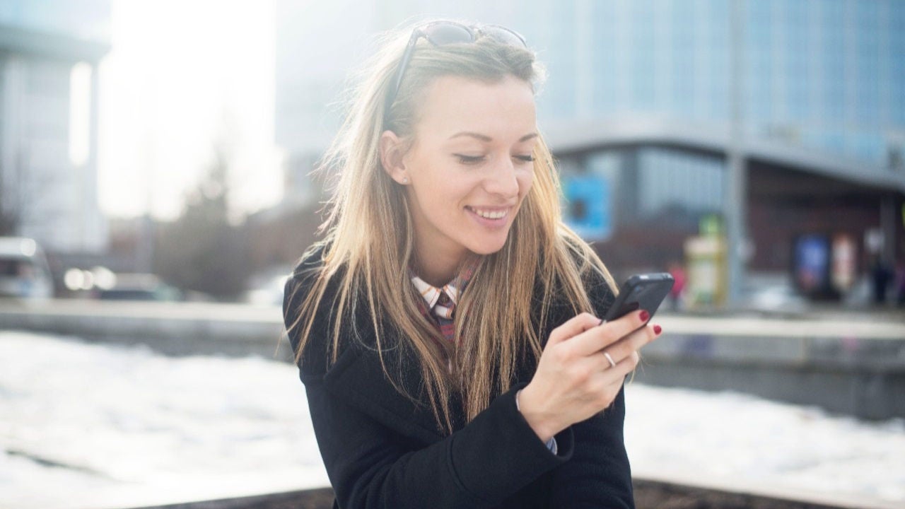 A younger woman sitting outside next to an office building looks at her smartphone.