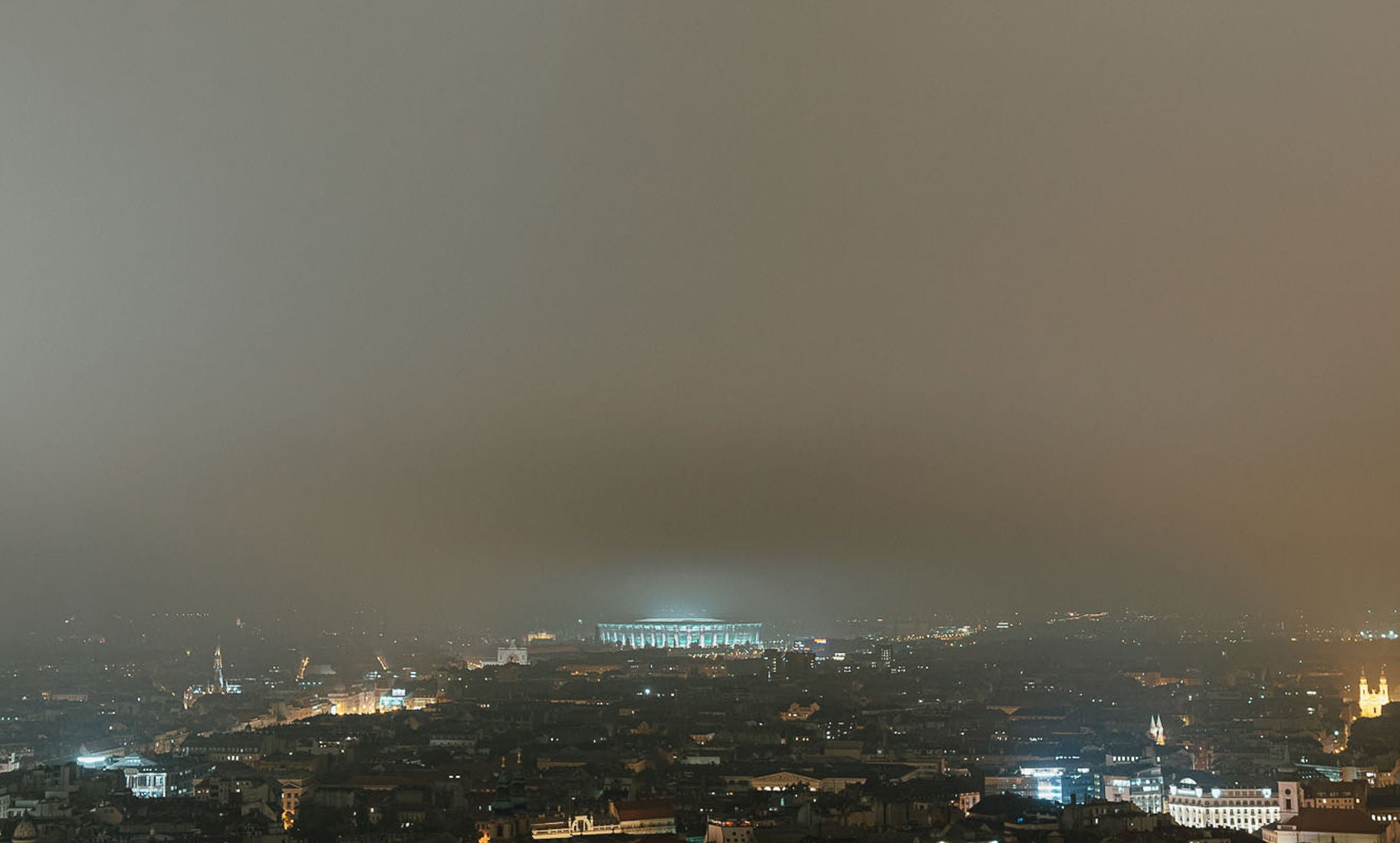 Éjszakai városkép ködös, narancsos árnyalatú égbolttal: a sötétben kivilágított város felett egy nagy, fényárban úszó stadion rajzolódik ki a horizonton, körülötte elszórtan világító utcák és épületek.