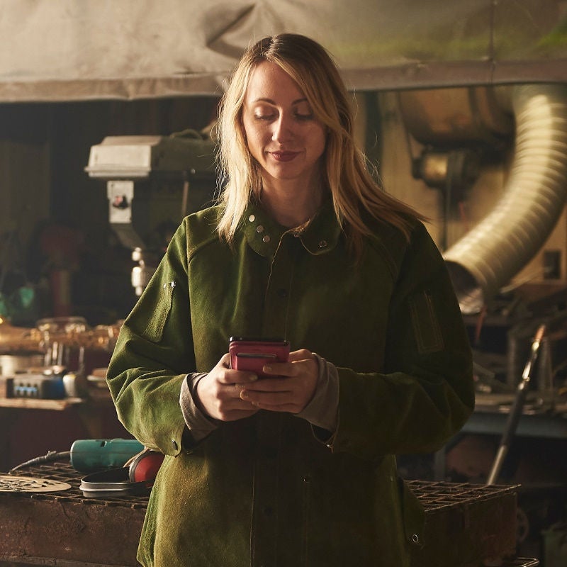 A woman in a heavy work coat looks at her phone in a workshop. 