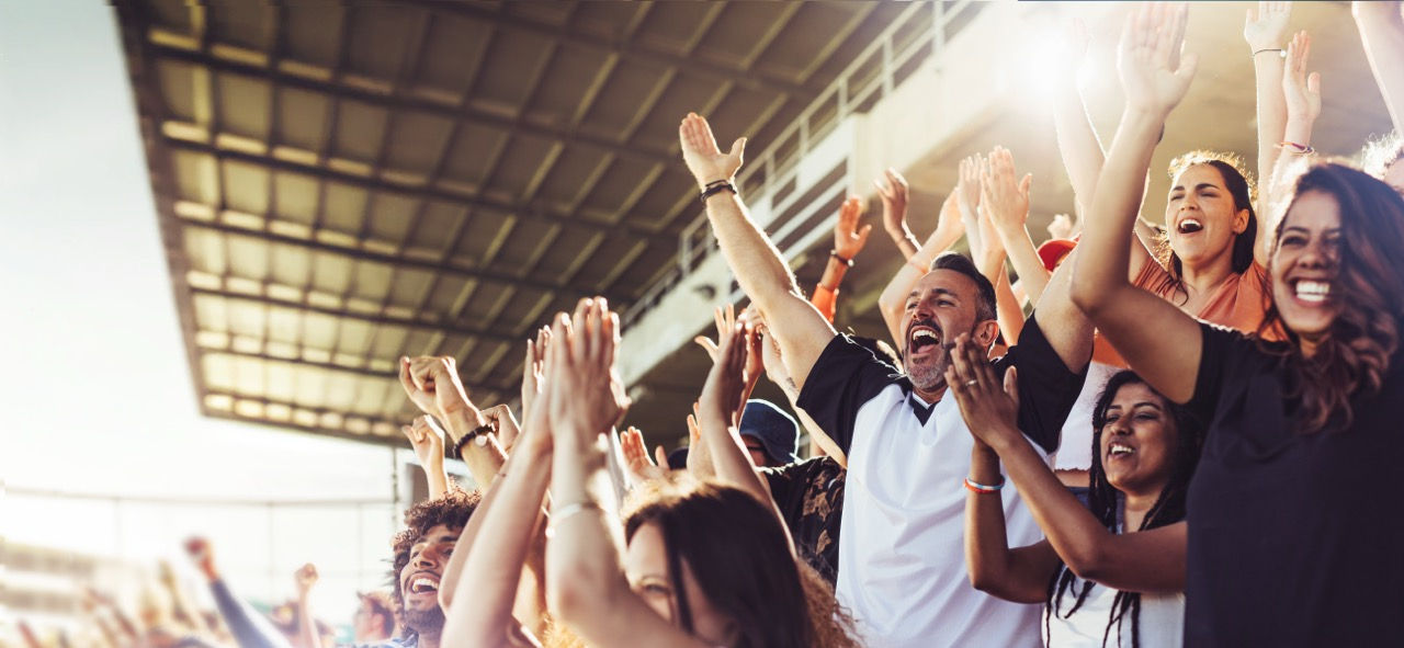 Crowd of sports fans cheering during a match in stadium. Excited people standing with their arms raised, clapping, and yelling to encourage their team to win.