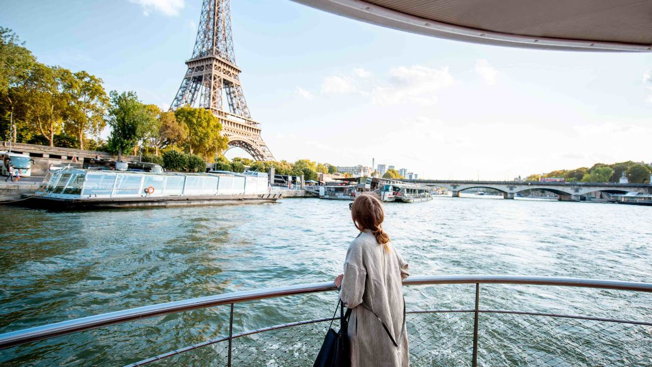 A woman in a coat stands at the railing of a boat as it passes the Eiffel Tower. 