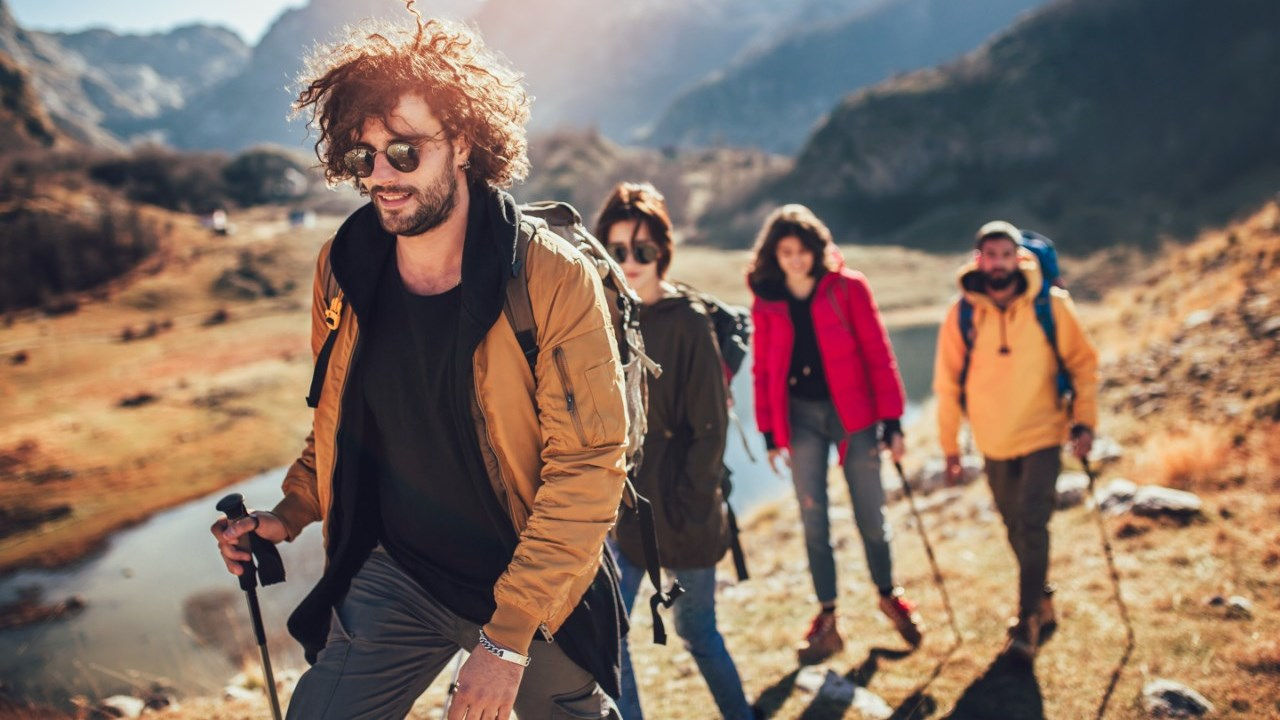 A group of young people in hiking gear and sticks walk along rugged terrain next to a river. 