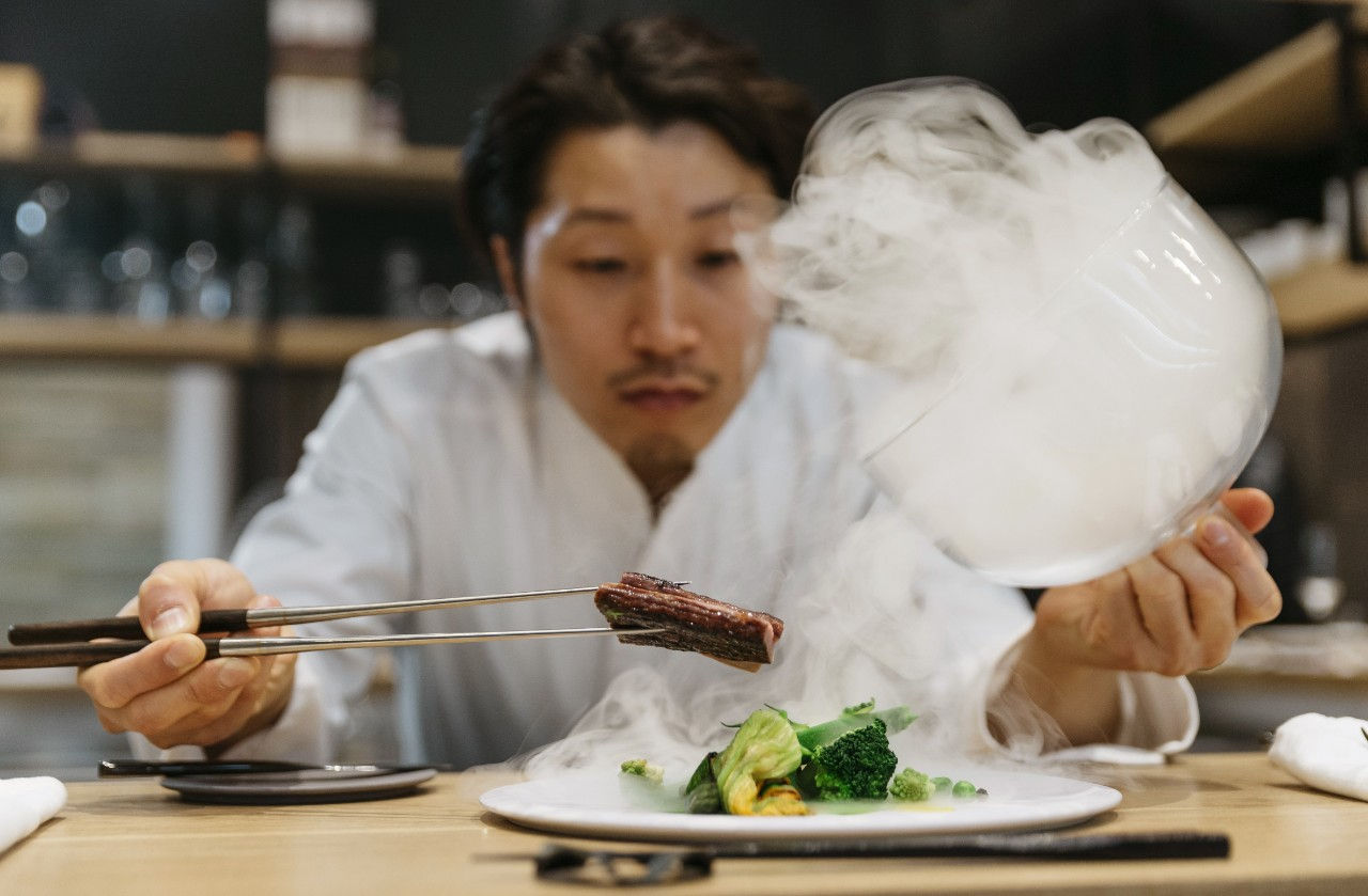 A chef holds a piece of meat over a dish while he lifts a cloque filled with steam. 