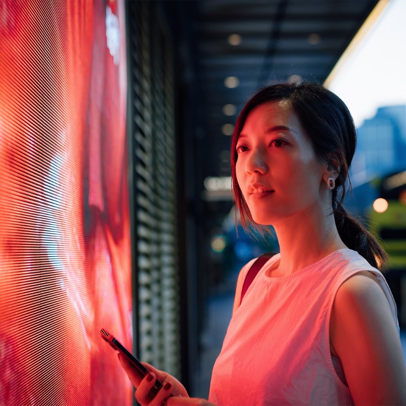 A young woman holds a smartphone and looks at a digital messaging board on a city street. 