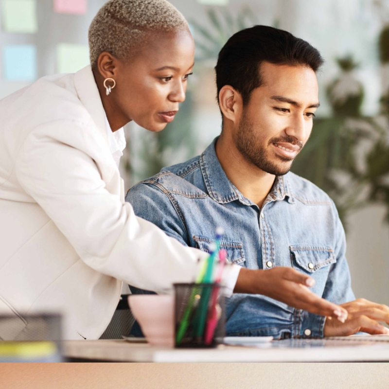 An office worker gestures to a laptop where a co-worker is seated. 