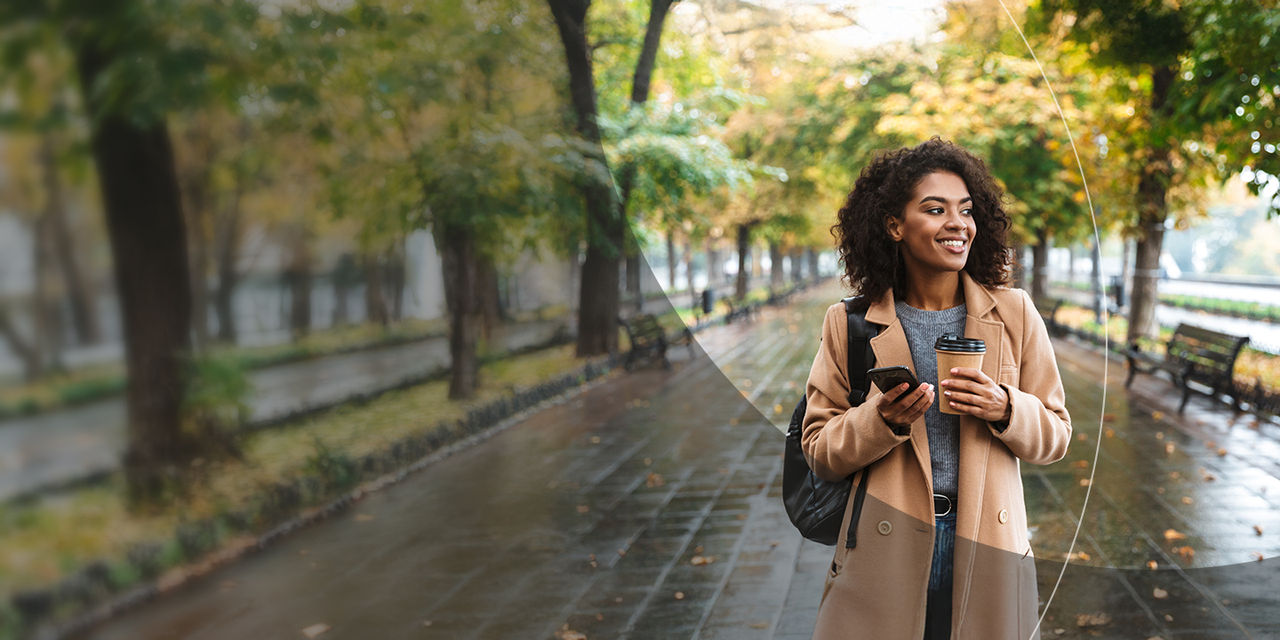 Une femme tenant un café et un smartphone tout en marchant dans un parc.