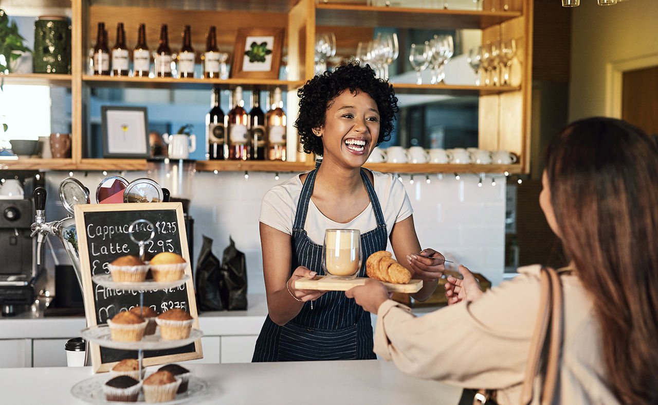 In een koffietentje geeft de ene vrouw een kopje koffie door aan de andere vrouw, waardoor een warme en vriendelijke sfeer ontstaat.