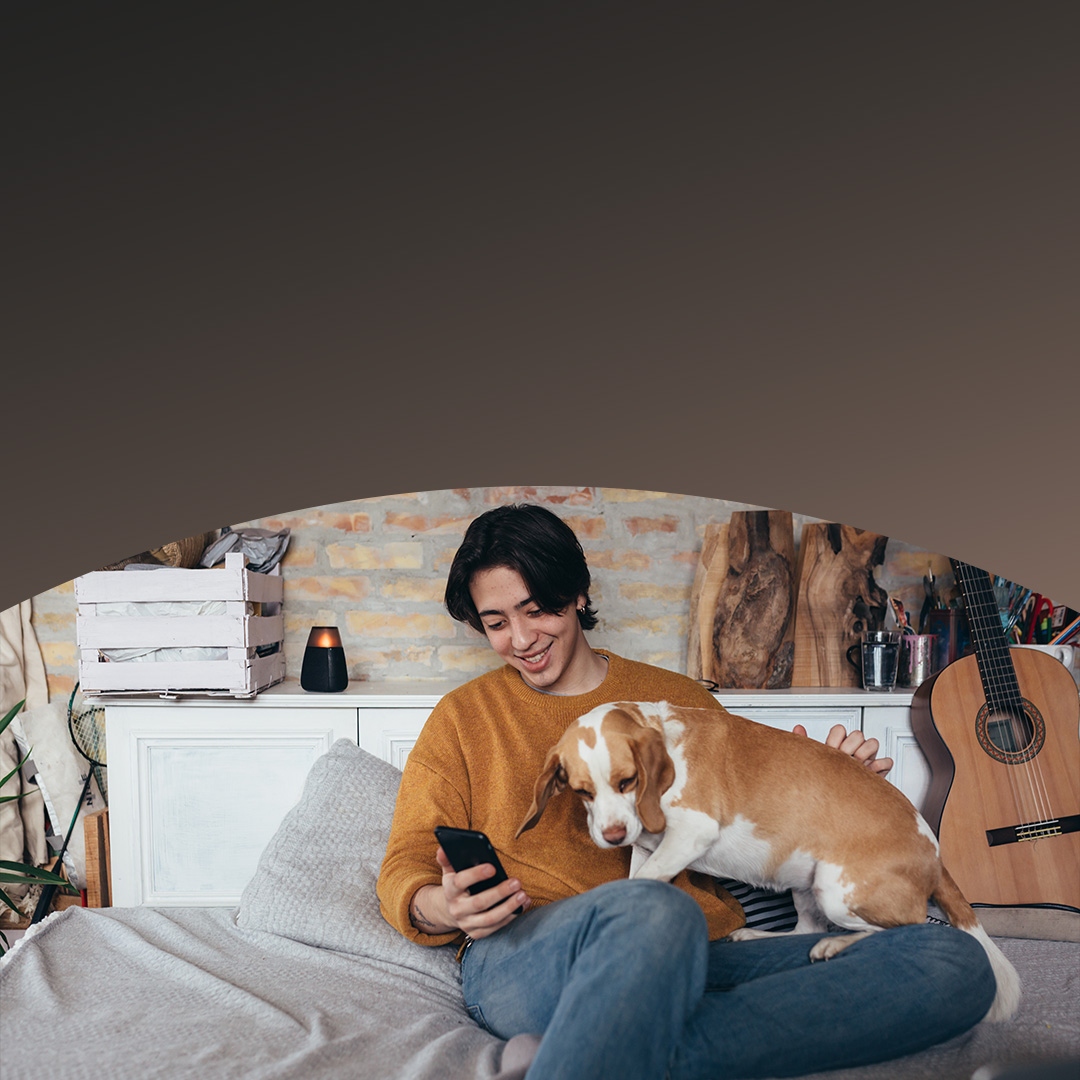 Een man en zijn hond zitten op een bed, waarbij de man een mobiele telefoon in zijn hand houdt en beiden er ontspannen uitzien.