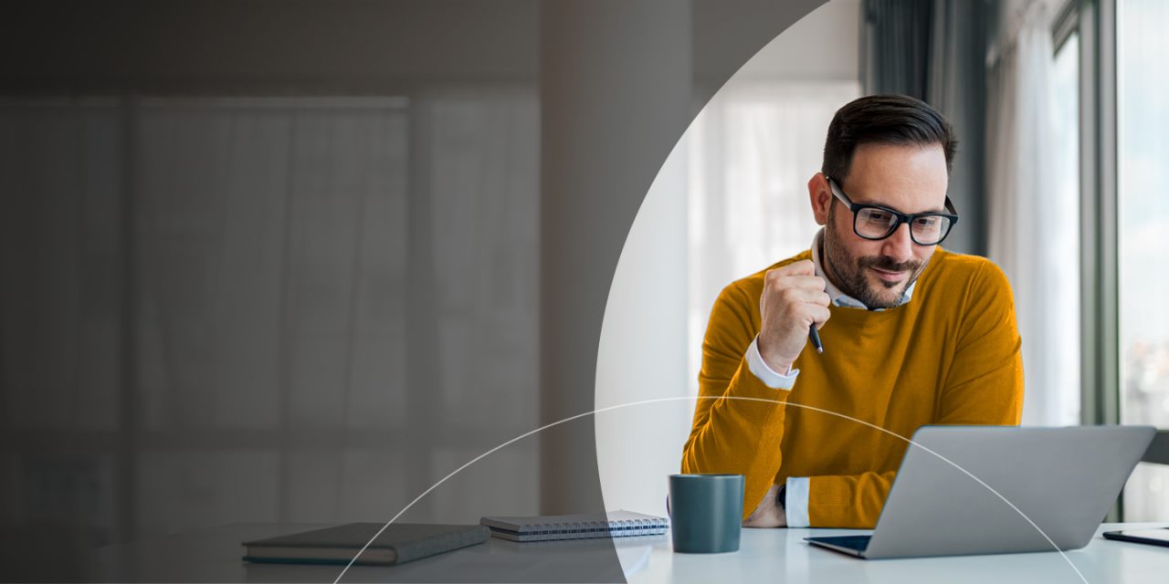 Мъж държи химикалка и гледа лаптоп.