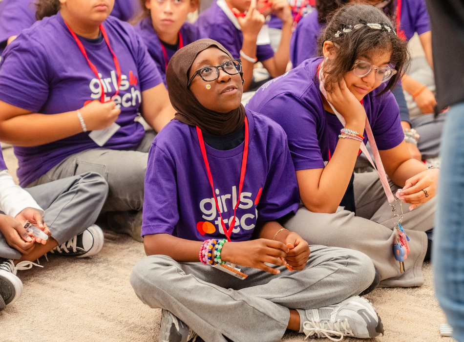 Uma garota vestindo um uniforme da Girls4Tech olha para uma instrutora durante um exercício de segurança cibernética. 
