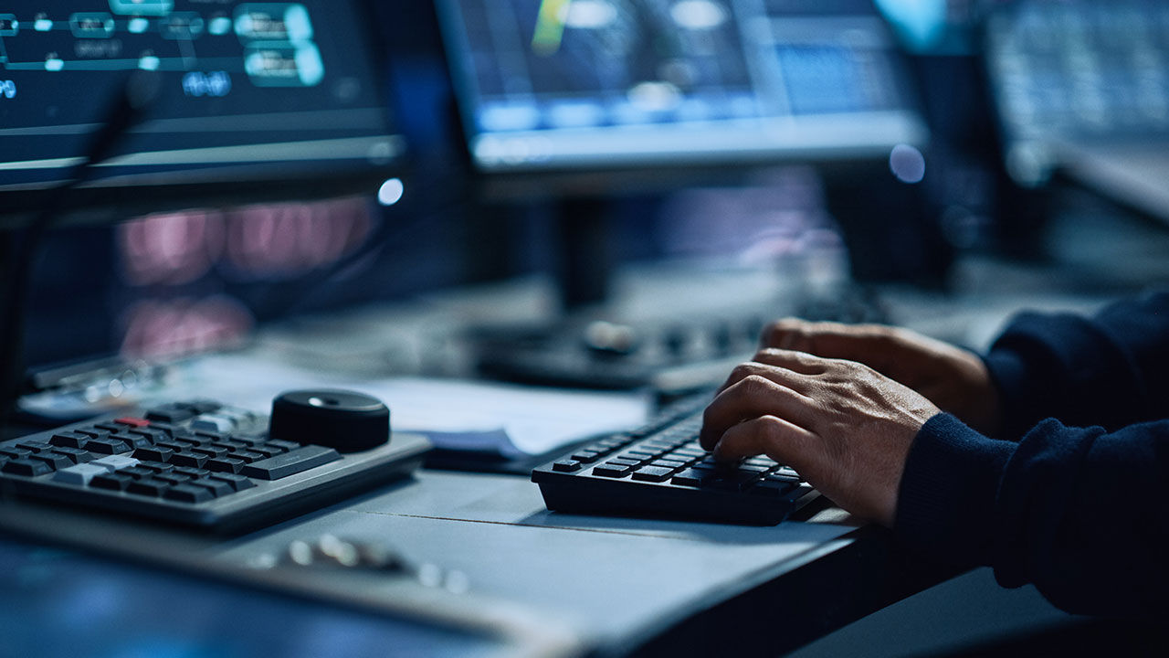 Especialista trabajando en computadora en sala de control de monitoreo tecnológico con pantallas digitales.