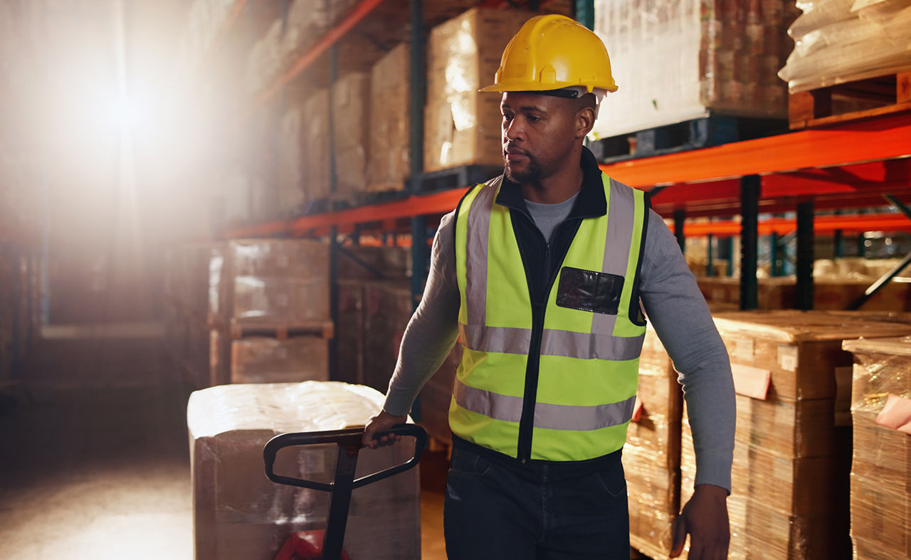 A man wearing a safety vest and hard hat walks through a warehouse, surrounded by shelves and equipment.