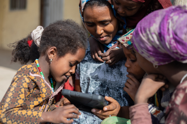Women and a girl gather around a tablet. 