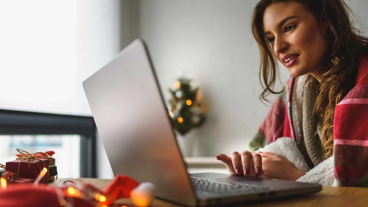 A woman looks at her laptop on a desk in a room decorated with holiday lights. 