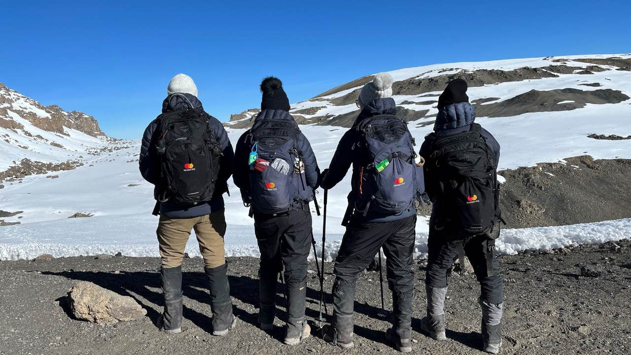 Four Mastercard employees in hiking gear showing off their Mastercard backpacks near the summit of Mount Kilimanjaro. 