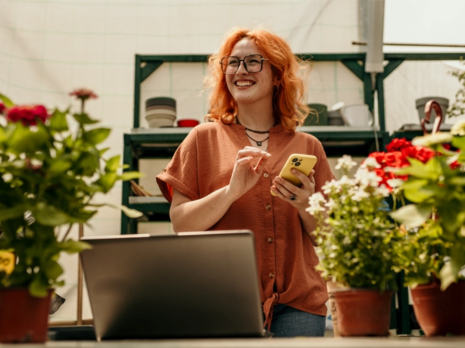 A woman holds a phone and looks over a laptop in a garden shop. 