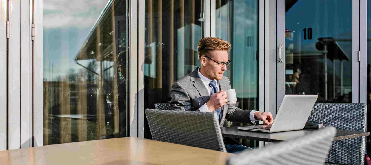 A businessman in a suit drinks coffee at an outdoor table in front of  a laptop. 