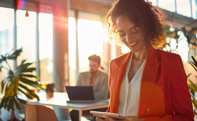 Une femme dans un bureau, concentrée sur sa tablette, assise à un bureau avec des papiers et une plante à proximité.