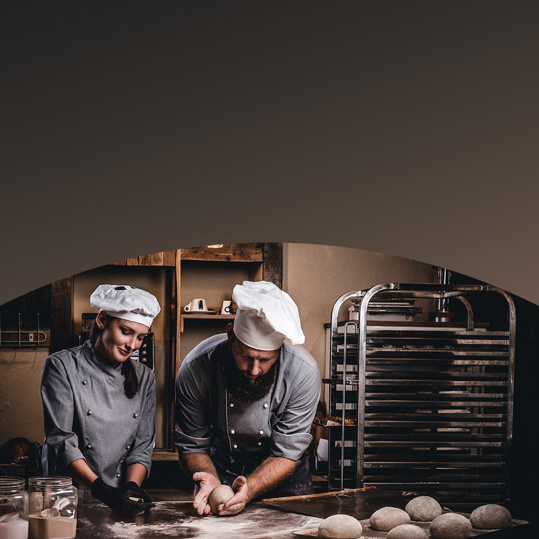 Dos chefs en la cocina de una panadería preparando e inspeccionando bolas de masa en el mostrador.