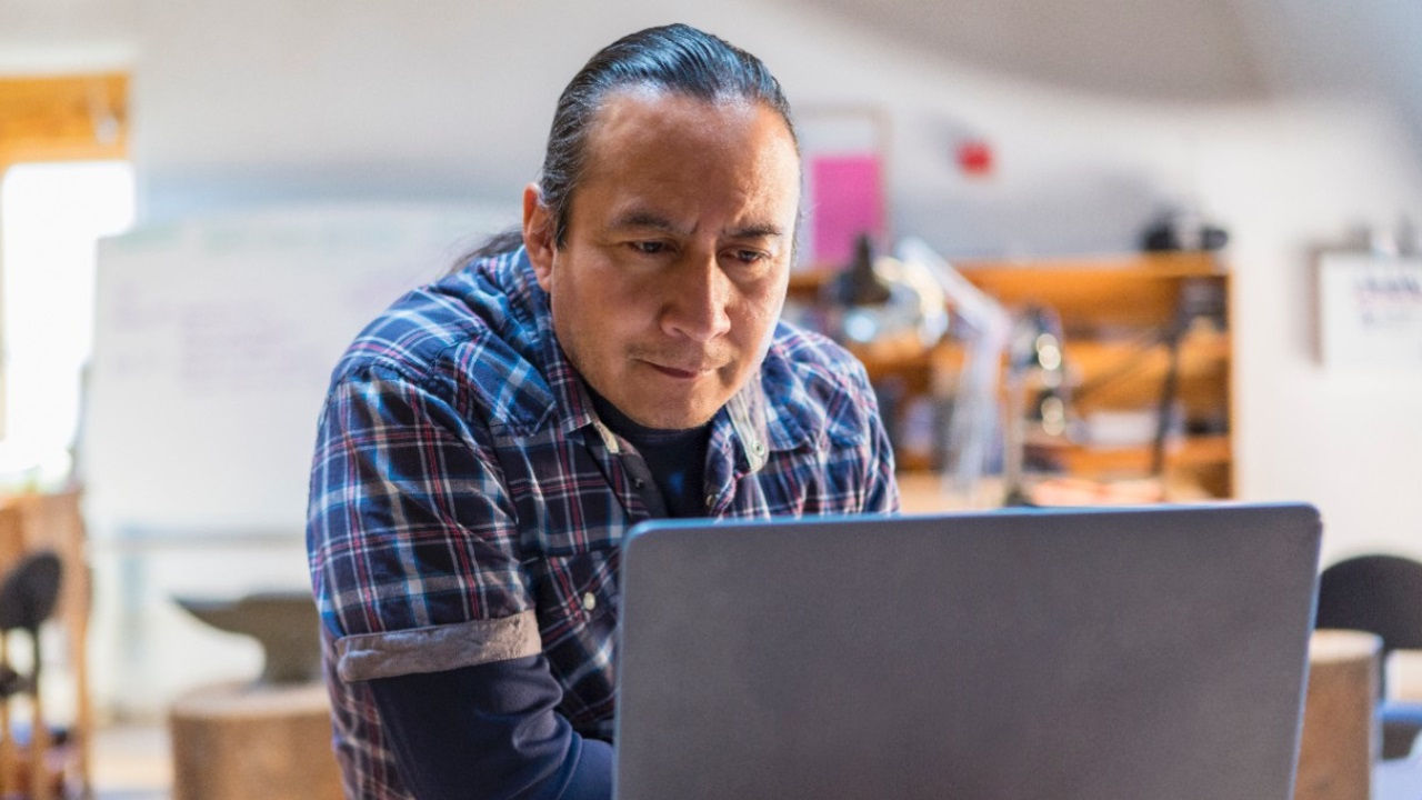 A man in a plaid shirt in a workshop looks at a computer. 