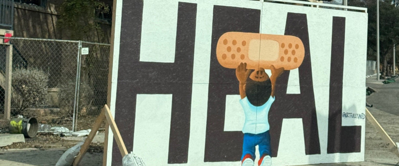 Un letrero que dice "HEAL" con un niño pequeño sosteniendo una curita sobre la E y la A, frente a la destrucción de incendios forestales. 