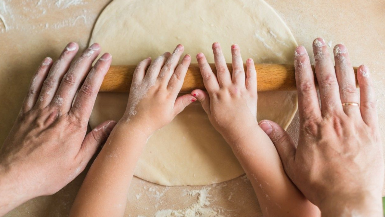 Two pairs of hands, one older, one child, roll out dough with a rolling pin. 