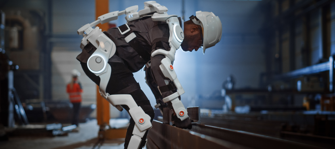 A man wearing an exoskeleten bends over to lift something in  an industrial building site.