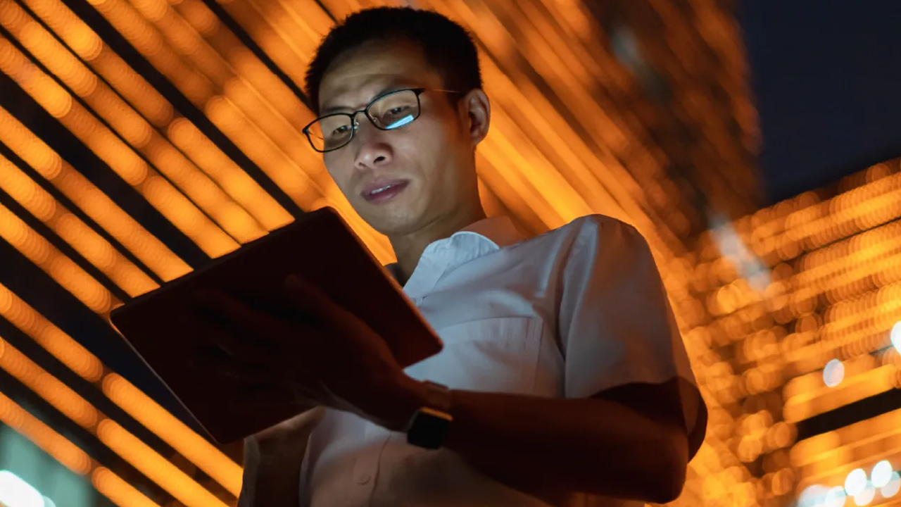 Man using a tablet under bright orange city lights at night.