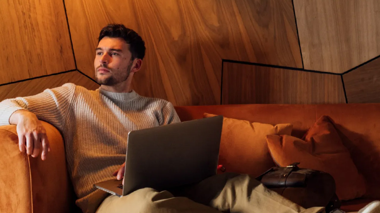 Man with a laptop sitting on a sofa in a wood-paneled room.