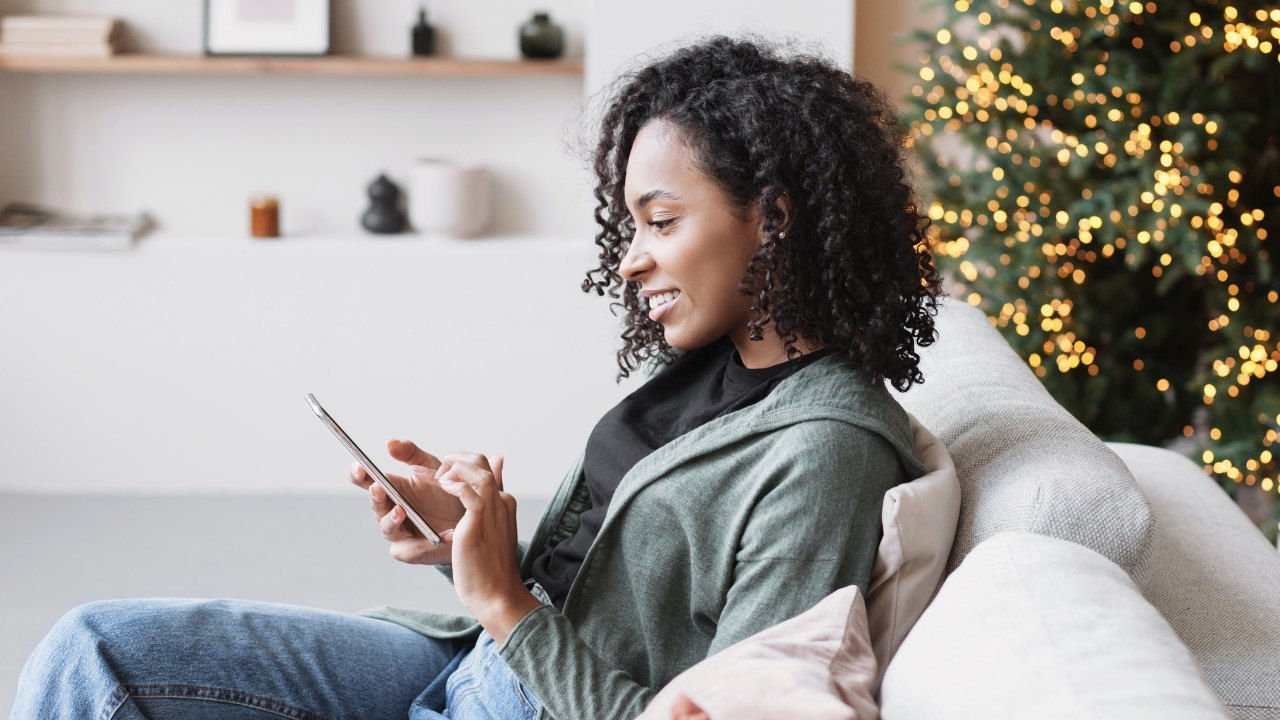 A woman sits on a couch looking at her phone in front of a decorated Christmas tree. 