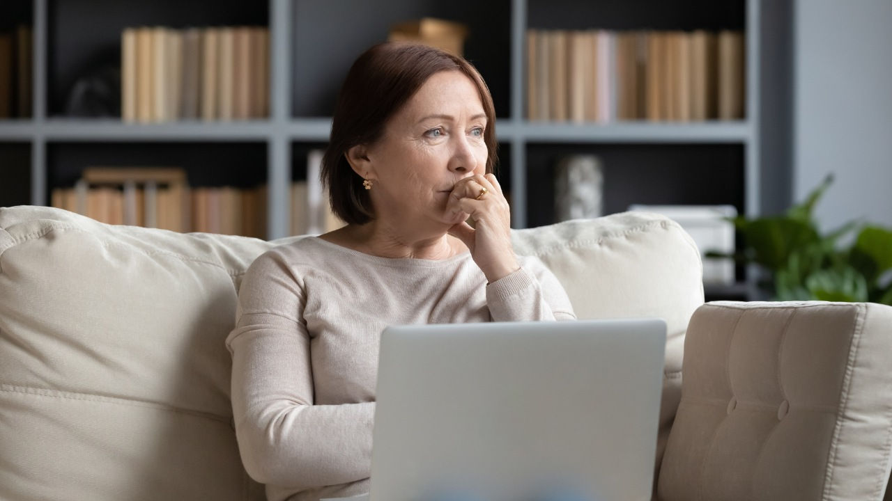 A middle-aged woman looks concerned  as she stares into the distance with a laptop on her lap while sitting on a couch. 