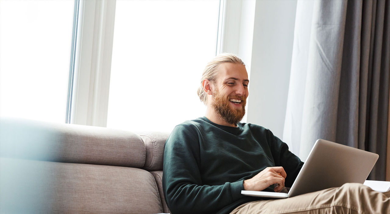 Mand, som smiler, mens han bruger sin laptop i sofaen