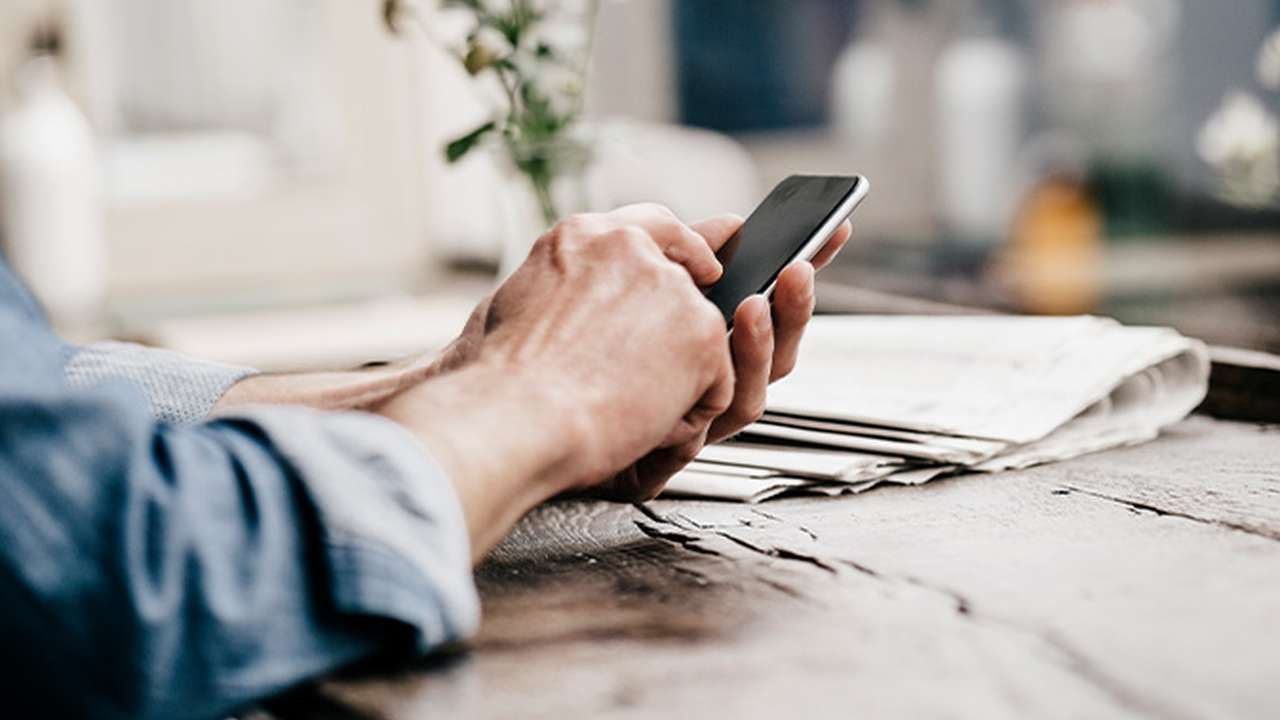 Hænder, der holder og bruger en mobiltelefon på et træbord.